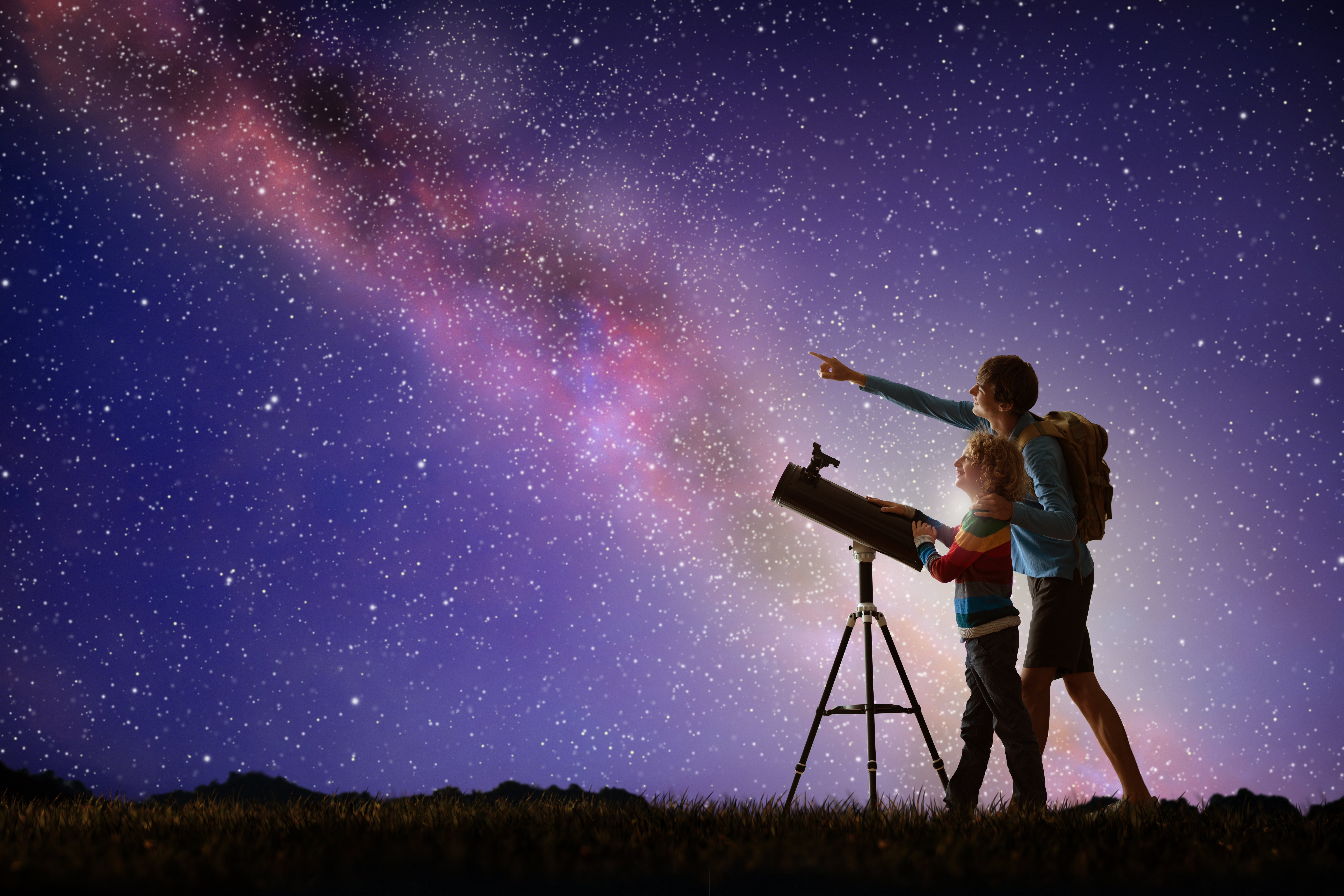 An adult points at the Milky Way while a child looks through a telescope on a grassy hill under a star-filled night sky.