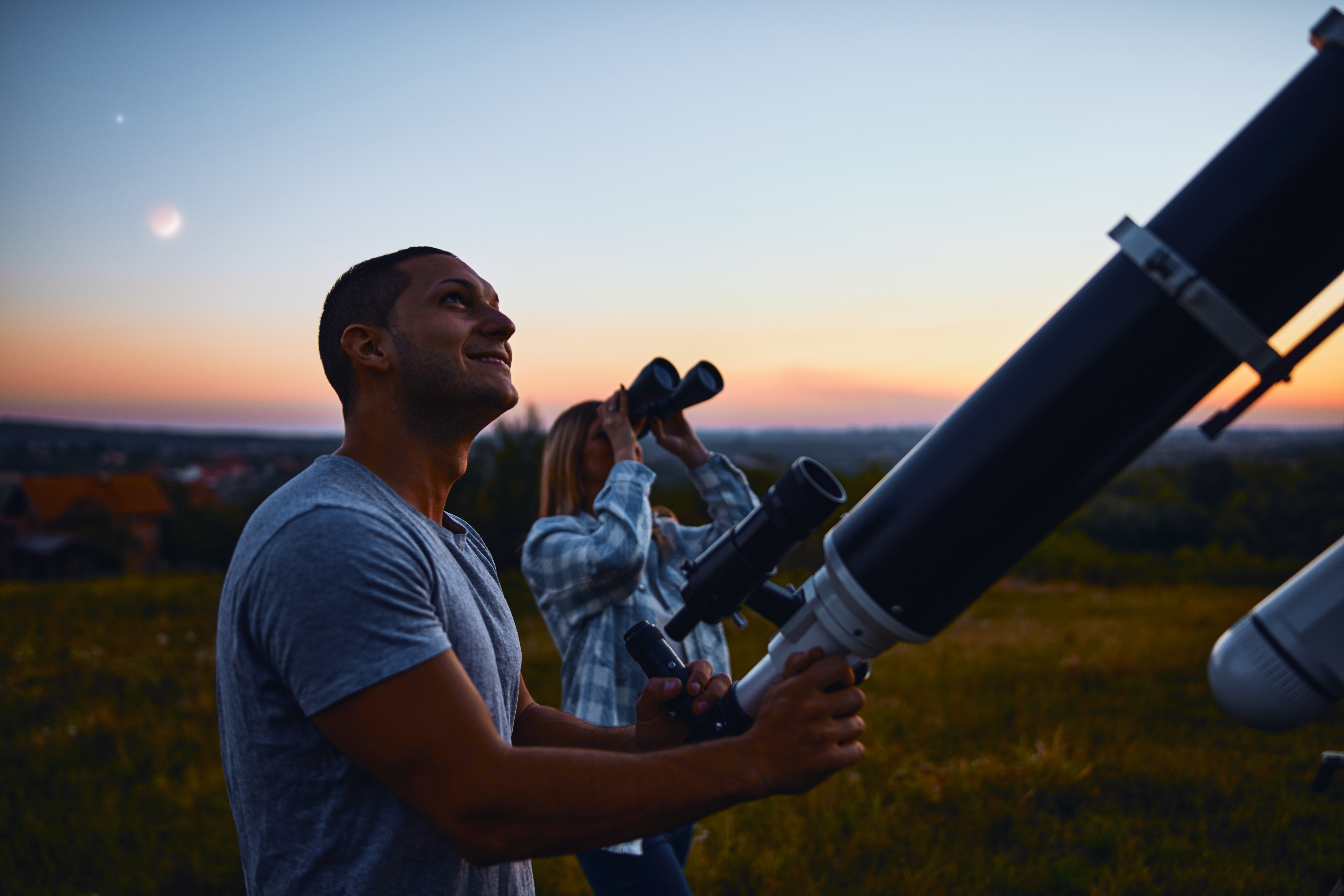 A man smiles while holding a telescope, and a woman gazes through binoculars under a twilight sky with a crescent moon.
