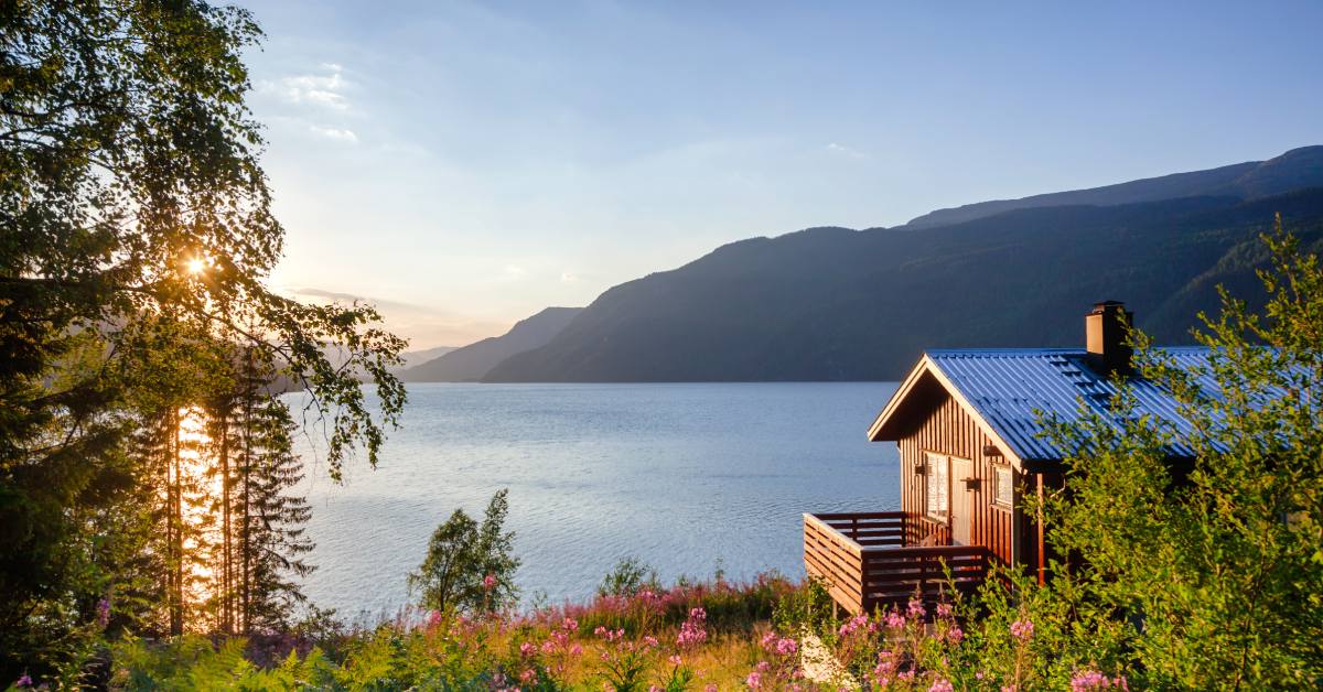 A wooden cabin with a metal roof overlooking a calm mountain lake at sunset, surrounded by wildflowers, trees, and mountains.