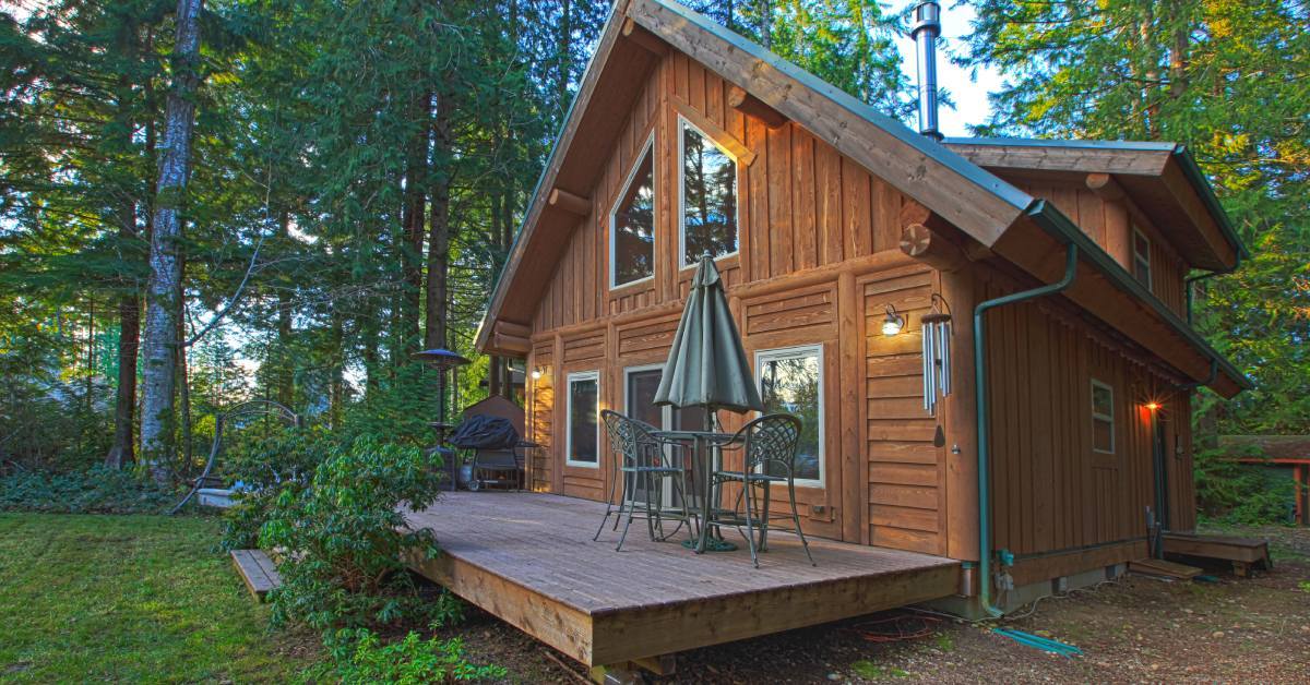 A rustic wooden cabin with an outdoor deck and a patio table, surrounded by tall pine trees in a forest setting.