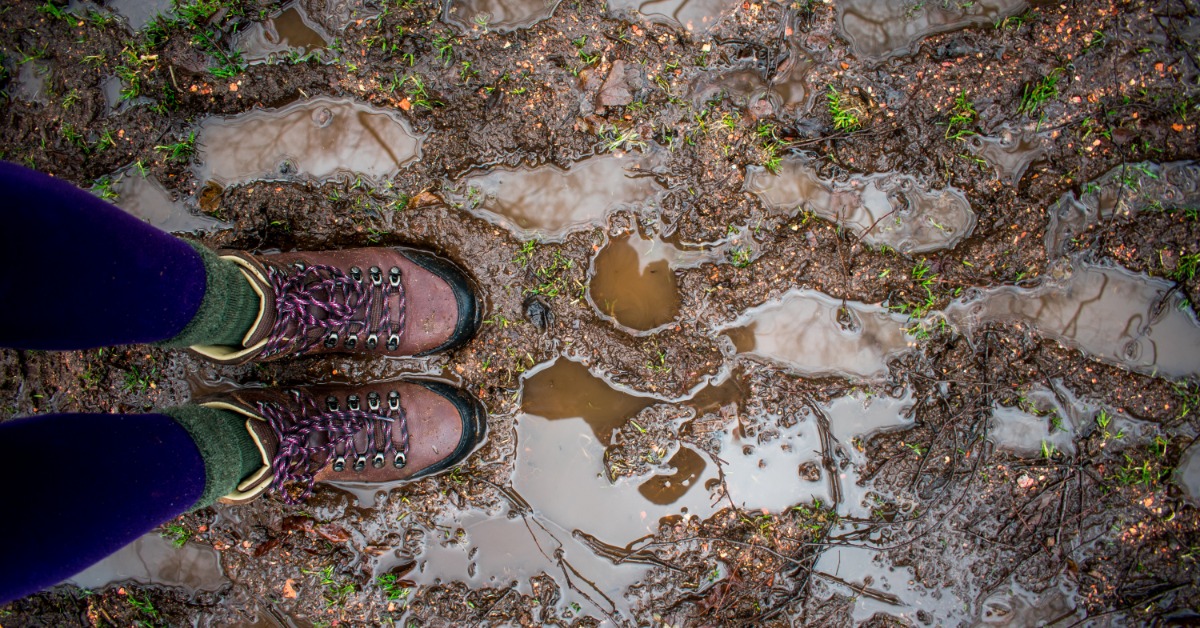 A person in brown hiking boots standing on a muddy trail with puddles, wet soil, and scattered grass.