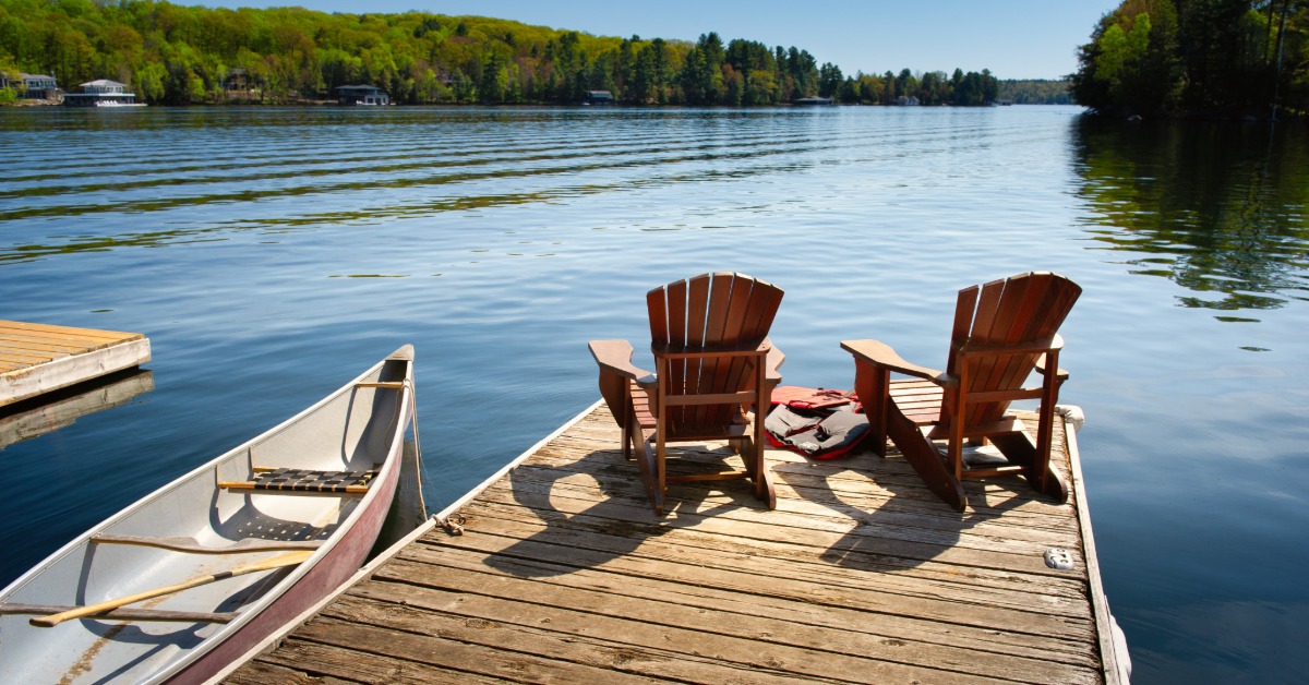 A wooden dock with Adirondack chairs and a canoe beside a calm lake surrounded by green trees on a sunny day.