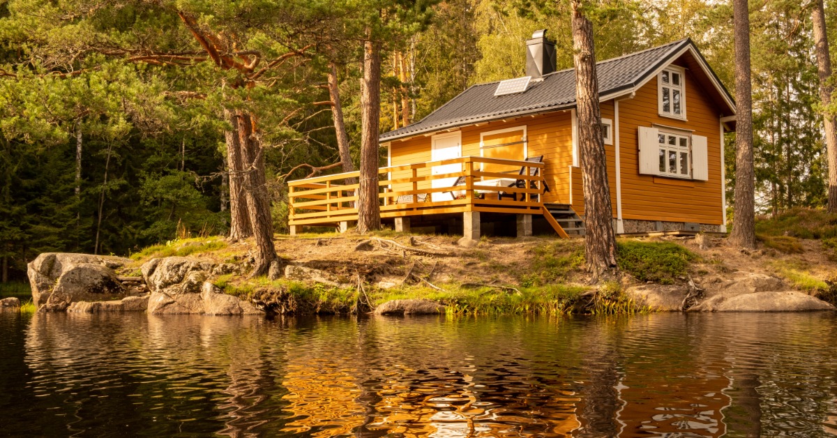 A small lakeside cabin surrounded by tall pine trees with warm sunlight reflecting across calm water in the foreground.