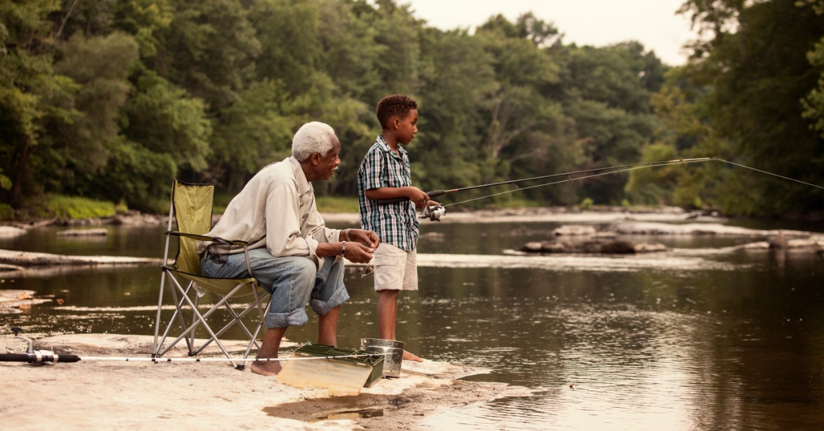 Grandfather and grandson fish together along a quiet riverbank, casting lines into calm water surrounded by trees.