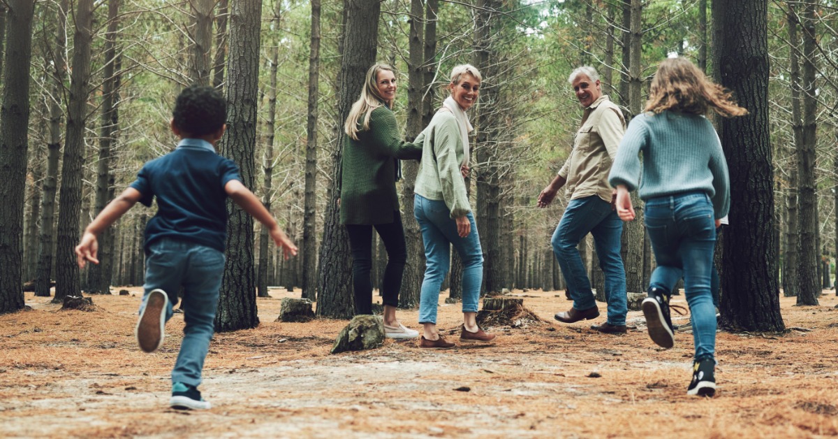 A family walks through a pine forest trail as children run ahead, with parents and grandparents smiling back.