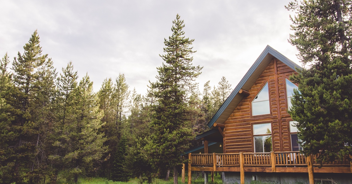 Large log cabin with a wraparound porch surrounded by tall pine trees and a grassy clearing beneath a cloudy sky.