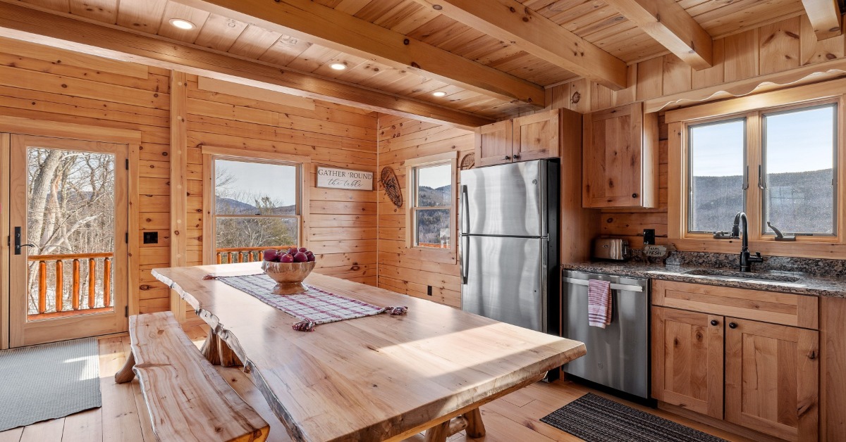 Rustic wood cabin kitchen with a large dining table, stainless steel appliances, and mountain views through the windows.