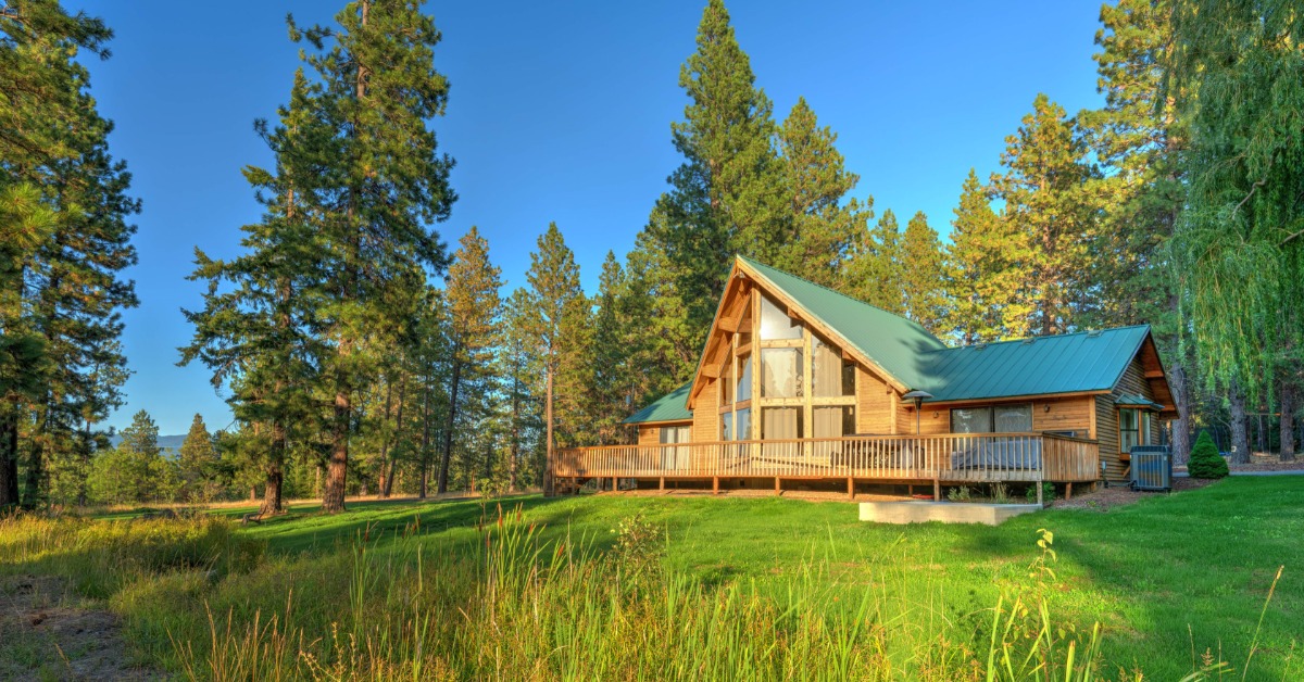 Spacious log cabin with a green metal roof and wraparound deck on a grassy lawn surrounded by tall pine trees.