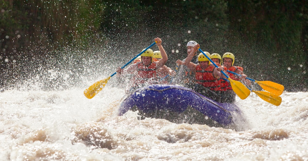 Rafters in yellow helmets paddle a purple raft through churning whitewater as waves splash high around the group.