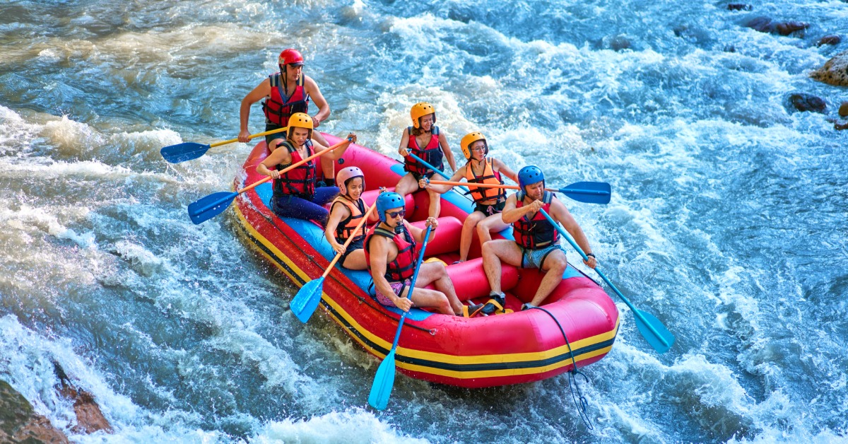 Group of rafters wearing helmets and life jackets paddle a red raft through rushing whitewater rapids on a rocky river.